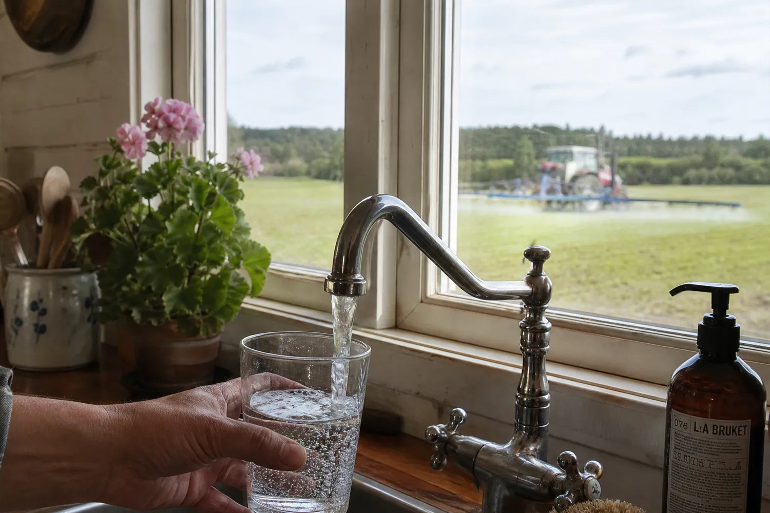 fyller glas med vatten, utanför besprutar traktor ett fält